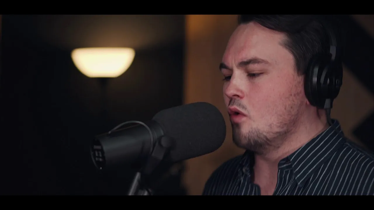 Man wearing headphones singing into a studio microphone with a floor lamp in the background.