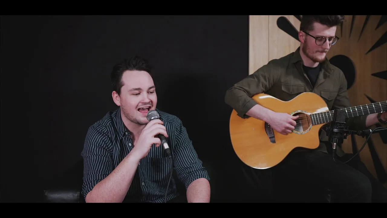 Man singing into a microphone while another man plays an acoustic guitar beside him.