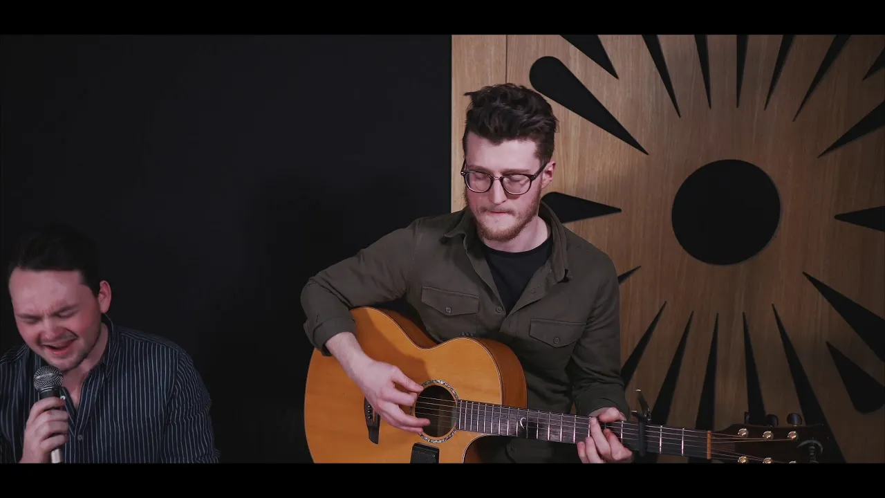 Two men performing music indoors; one singing into a microphone and the other playing an acoustic guitar.
